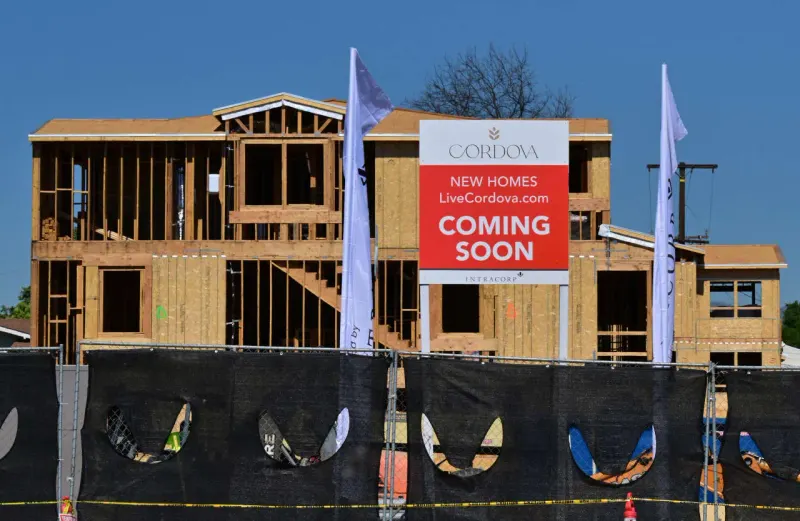 A small house is pinned beneath a rising metal spring, symbolizing mortgage-rate pressure on housing.