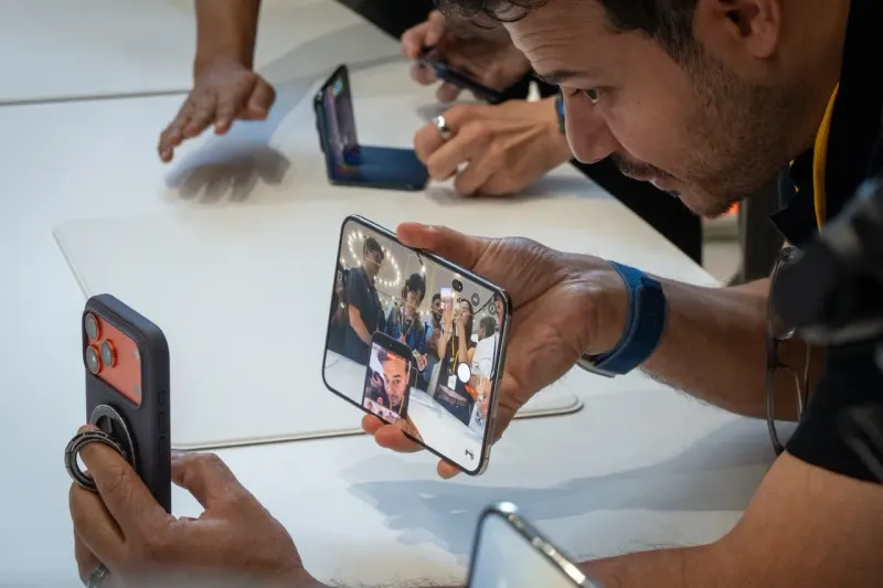 Three cables lower a folding smartphone from a tall stack of coins, symbolizing carrier efforts to cut its $1,999 cost.