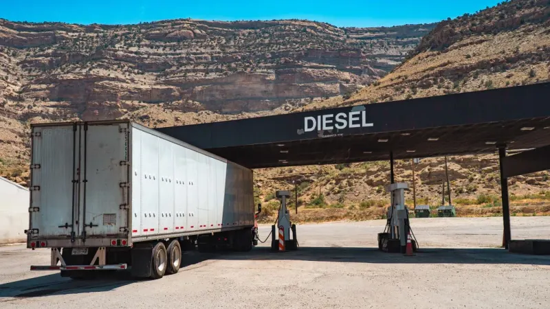 A heavy diesel droplet crushes an economic gear while a market arrow remains above the impact.