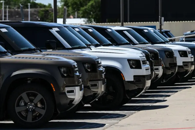 A factory gear rolls forward on new orders while flames and tariff barriers constrict its path.